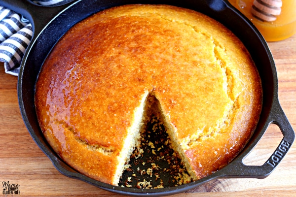 skillet of gluten-free cornbread with one slice cut out with honey and a kitchen towel in the background