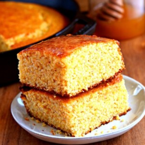 2 slices of gluten-free cornbread stacked on top of each other with a honey jar and a skillet of cornbread in the background