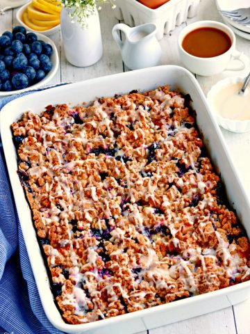 a pan of gluten-free blueberry breakfast casserole with blueberries, lemon, egg, flowers, coffee, cream and glaze in the background.