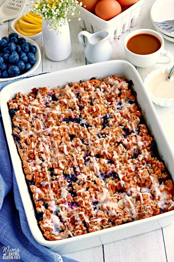 a pan of gluten-free blueberry breakfast casserole with blueberries, lemon, egg, flowers, coffee, cream and glaze in the background.