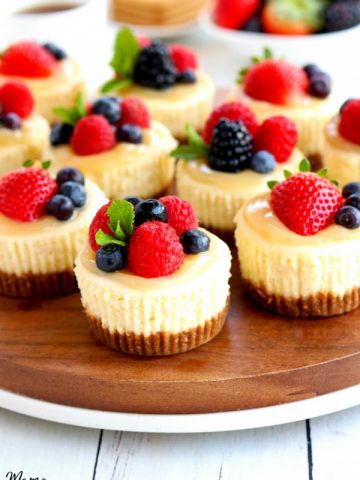 mini cheesecakes topped with berries on a wood platter with berries, graham crackers and coffee in the background