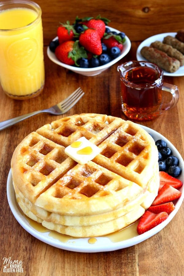 gluten-free waffles with butter and syrup on a white plate with berries. A fork, syrup, sausage, berries and a glass of orange juice is in the background.