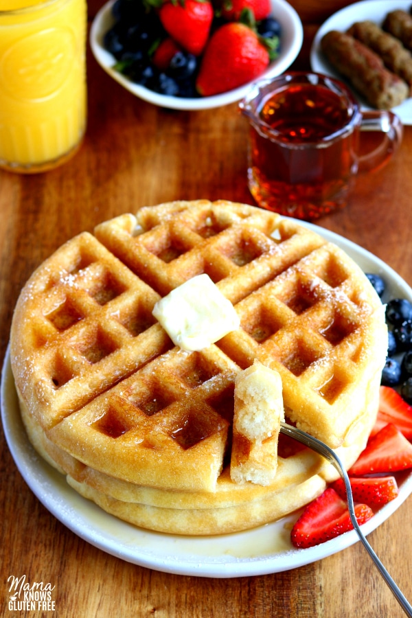 gluten-free waffles with a bite cutout of it on a fork with syrup, berries, sausage, and orange juice
