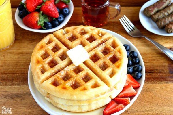 a plate of 3 gluten-free waffles with butter, syrup, a fork, berries, syrup, sausage and orange juice