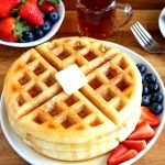 gluten-free waffles with butter and syrup on a white plate with berries. A fork, syrup and berries in the background.