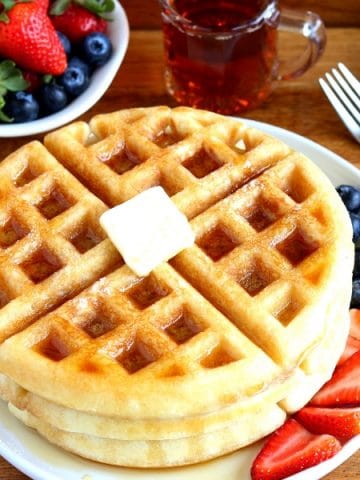 gluten-free waffles with butter and syrup on a white plate with berries. A fork, syrup and berries in the background.