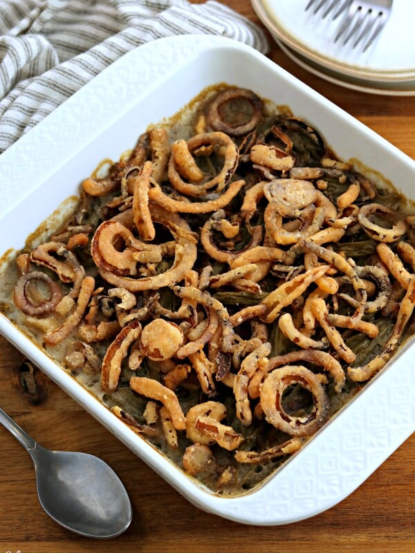 gluten-free green bean casserole in a white dish with a spoon, kitchen towel, forks and plates in the background