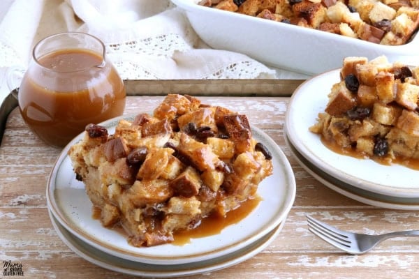 two servings of gluten-free bread pudding on white plates on a wooden tray with caramel sauce and baking dish in the background