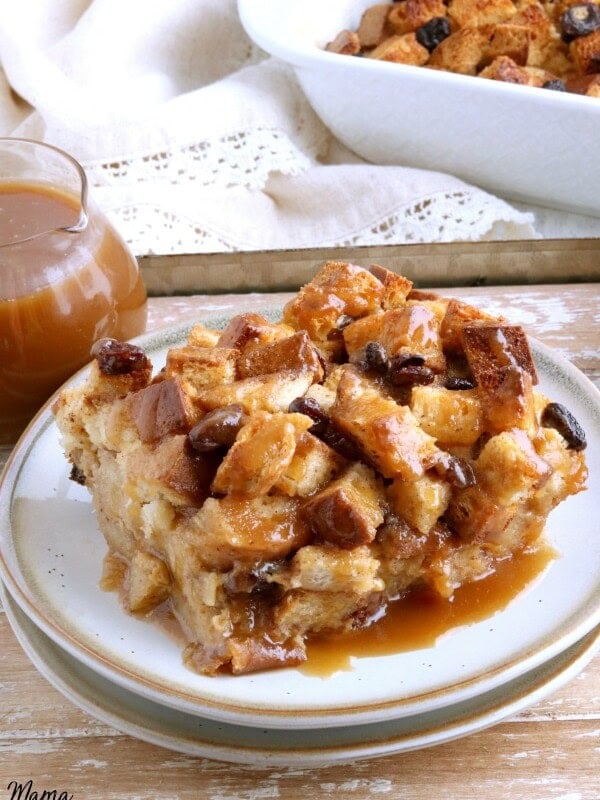 slice of gluten-free bread pudding on a white plate with caramel sauce and pan of bread pudding in the background