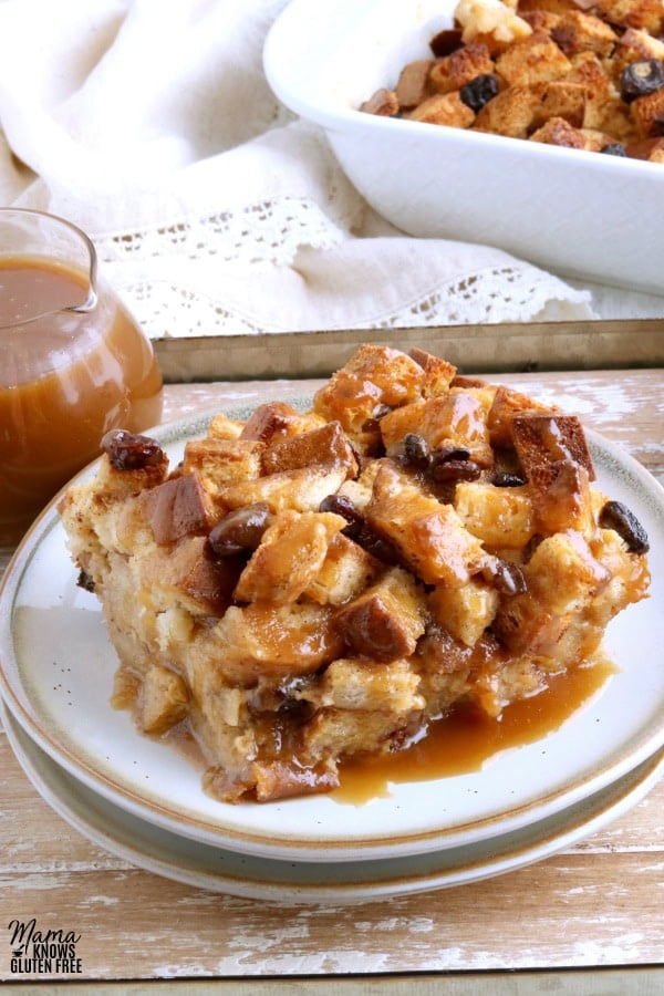 slice of gluten-free bread pudding on a white plate with caramel sauce and pan of bread pudding in the background