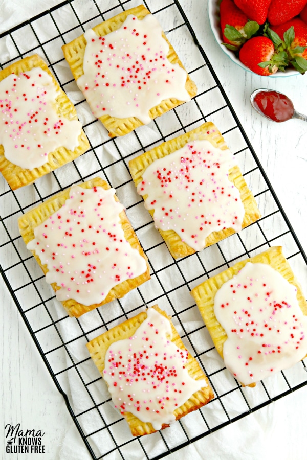 cooling rack of gluten-free pop tarts that are glazed, topped with sprinkles, and strawberries in a blue bowl.
