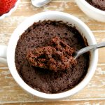 gluten-free chocolate mug cake with a spoon, another mug cake and strawberries in the background