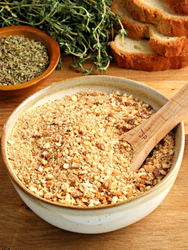 gluten-free bread crumbs in a bowl with a spoon and bread and herbs in the background