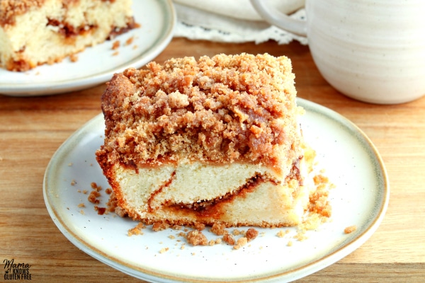 piece of gluten-free coffee cake on a white plate with a cofee cup and another slice of cake in the background