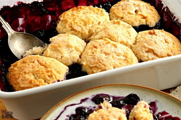 gluten-free blueberry cobbler in a white baking dish with a spoon and a serving on a plate