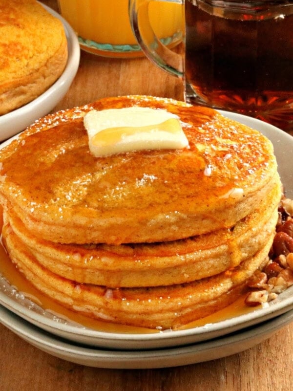 gluten-free pumpkin pancakes topped with butter and syrup on a white plate with more pancakes and syrup in the background