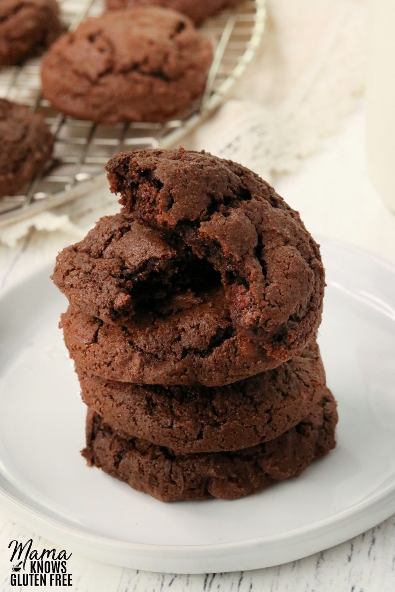 four gluten-free chocolate cookies stacked on a white plate with the cookies in teh background on a cooling rack