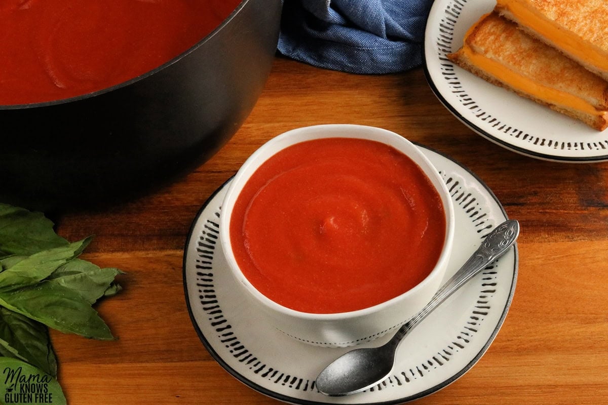 gluten-free tomato soup in a white bowl with grilled cheese sandwiches and the pot of soup, and fresh basil in the background