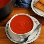 gluten-free tomato soup in a white bowl with a spoon, pot of soup and grilled cheese sandwich in the background