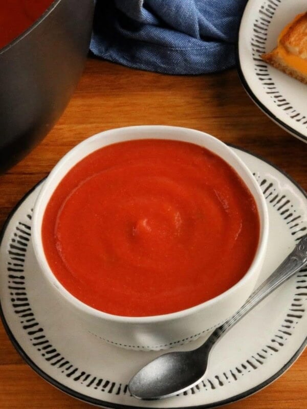 gluten-free tomato soup in a white bowl with a spoon, pot of soup and grilled cheese sandwich in the background