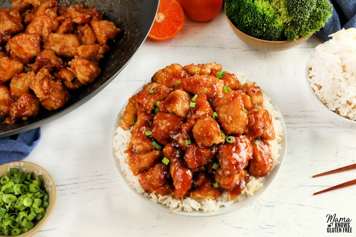 gluten-free orange chicken served over rice in a white bowl with rice, broccoli, oranges, green onions, chopsticks, and the wok of chicken in the background 
