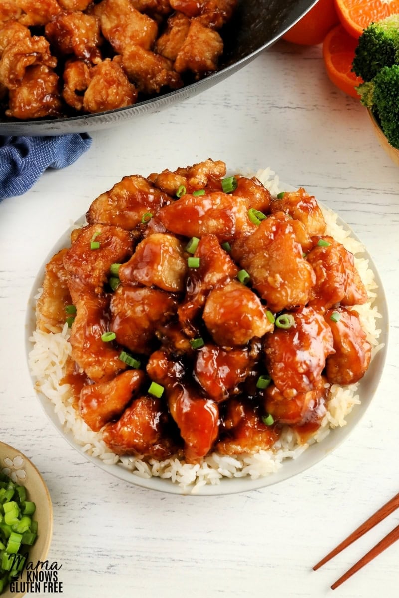 gluten-free orange chicken over rice in a white bowl with the wok of chicken in the background