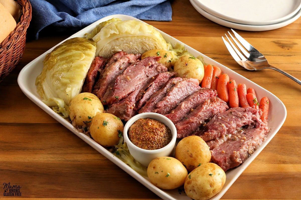 corned beef, cabbage, carrots, potatoes, and mustard on a white platter with plates, a fork and rolls in the background