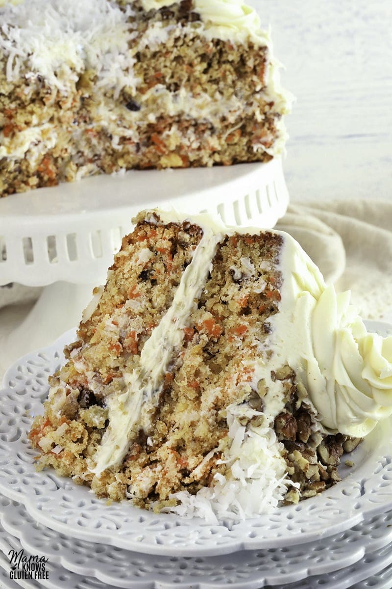 gluten-free carrot cake slice on a white plate with the cake on a white cake stand in the background