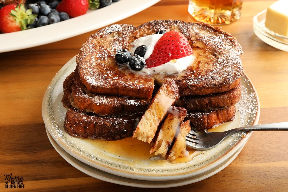 gluten-free French Toast topped with whipped cream, berries and syrup on a white plate with a fork with butter, syrup and platter of French toast and berries in the background