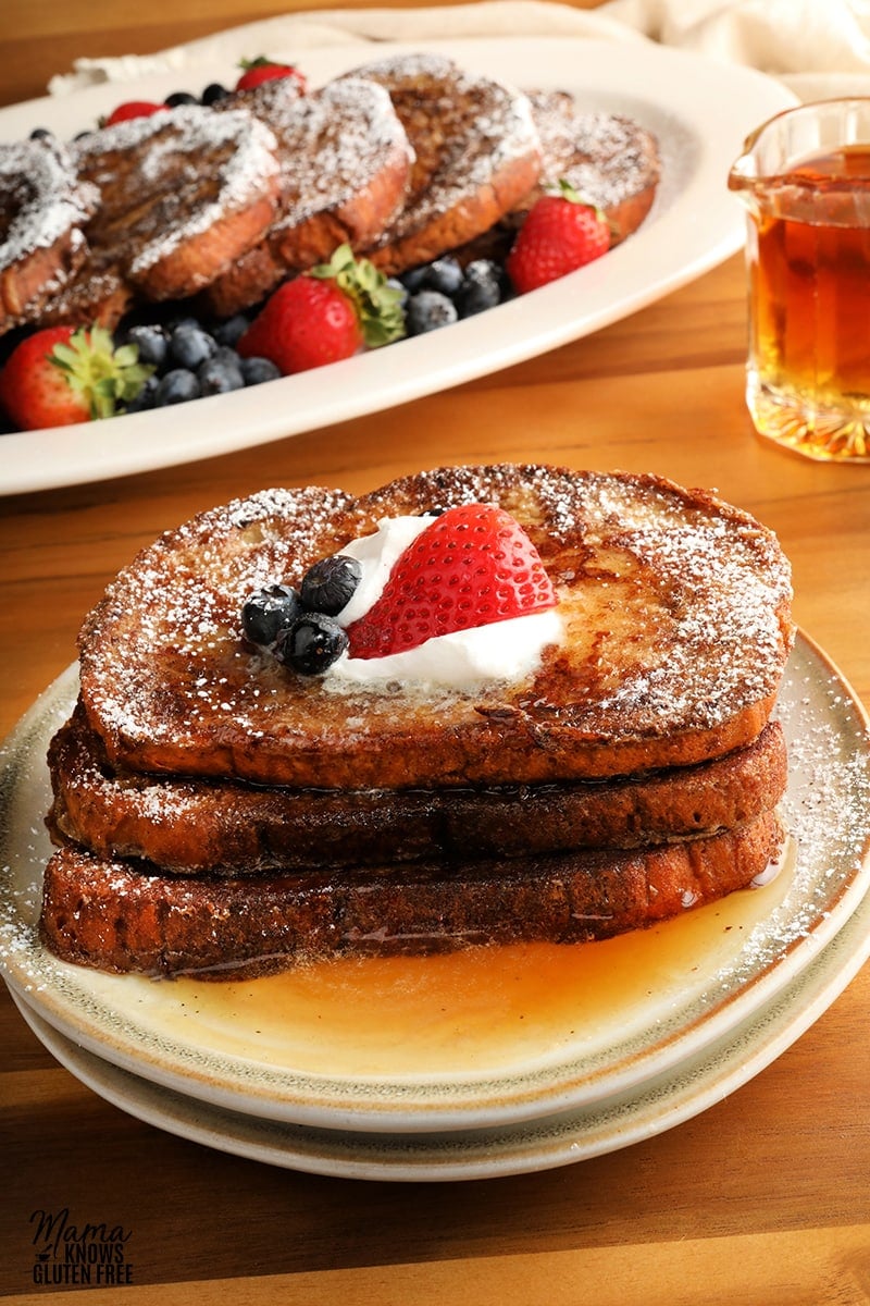 three pieces of gluten-free French Toast slices topped with berries, whipped cream, and syrup on a white plate with a platter for French Toast and syrup in the background