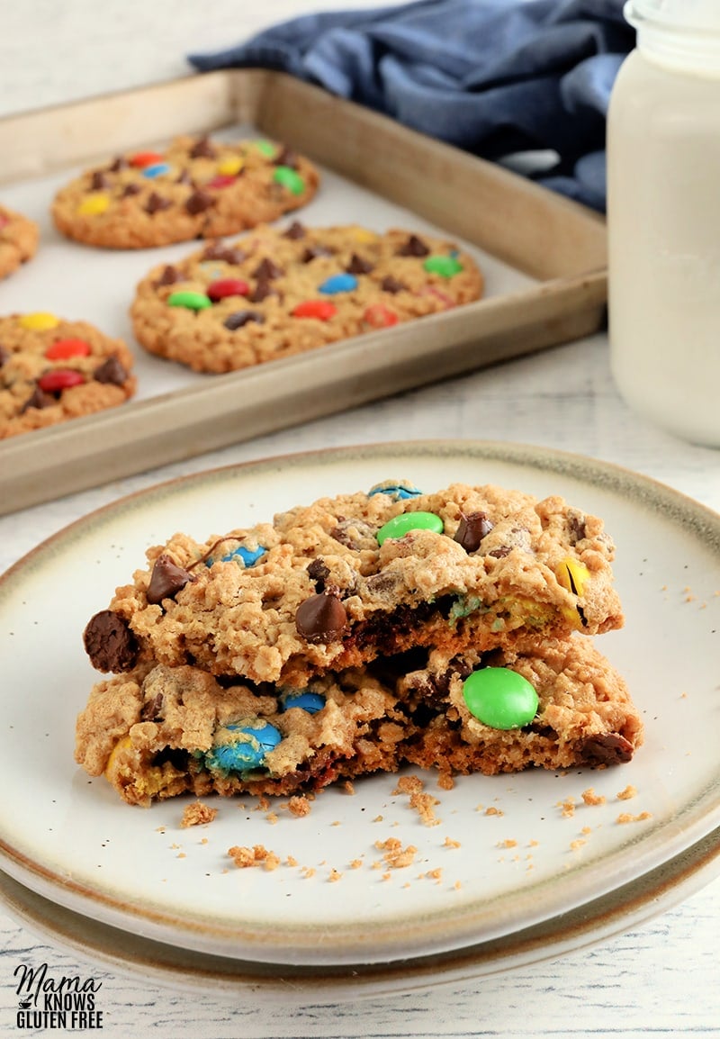 gluten-free monster cookie broken in half on a white plate with the cookies on a baking sheet in the background