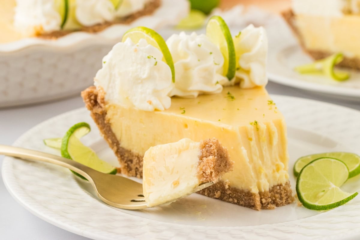 A close-up of a slice of gluten-free key lime pie on a white plate, with a bite taken out. A gold fork holds a piece of the creamy, tangy filling with a firm Chex crust. The pie is topped with whipped cream, lime slices, and a sprinkle of lime zest. The whole pie and additional slices are slightly blurred in the background.