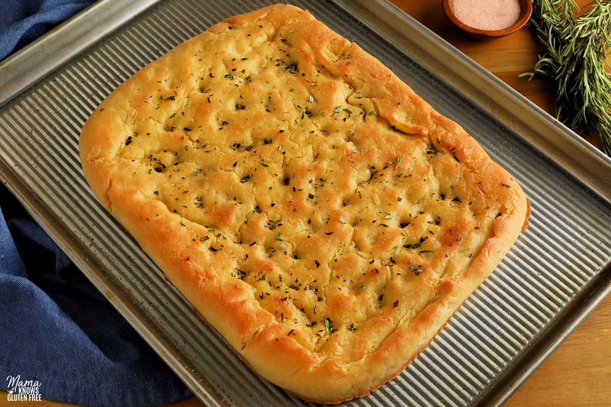gluten-free focaccia bread on a sliver baking pan with fresh rosemary and bowl of salt in the background