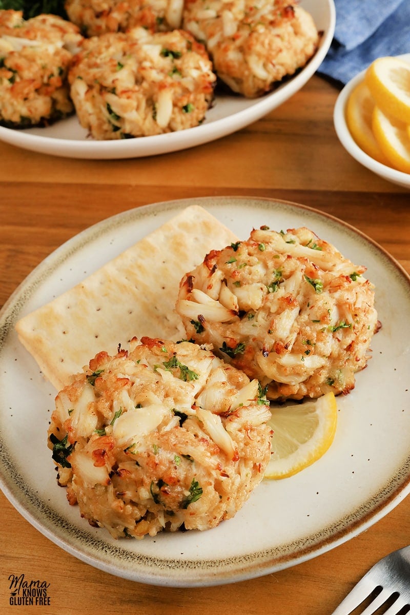 two gluten-free crab cakes on a white plate with a lemon slice and crackers with a platter of crab cakes and lemon slices in the background