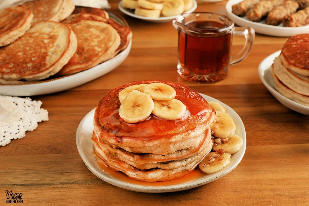 gluten-free banana pancakes on a white plate with syrup, bananas, sausage, and pancakes in the background