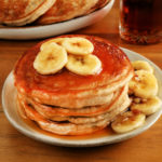 gluten-free banana pancakes on a white plate with syrup and pancakes in the background