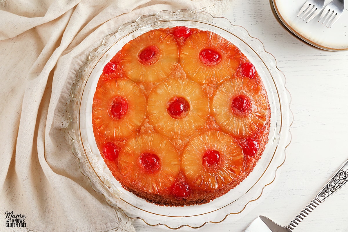 gluten-free pineapple upside down cake on a glass cake plate with a cake cutter and plates in the background