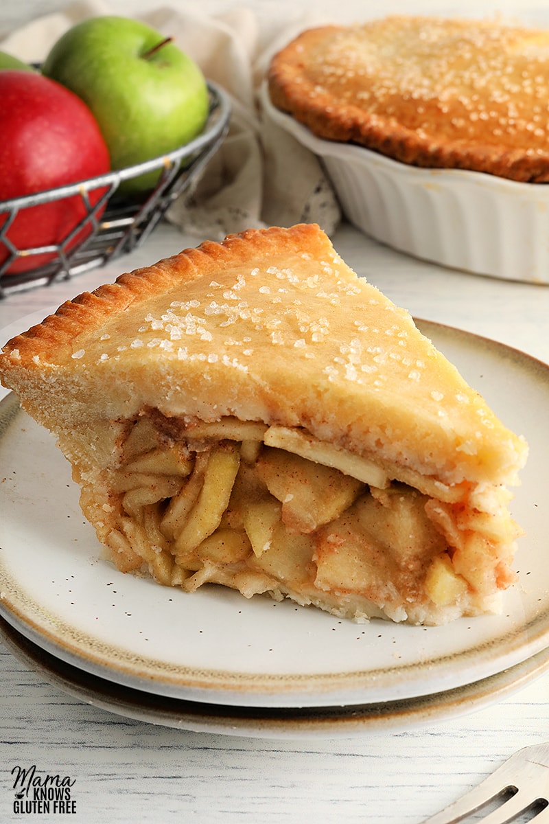 slice of gluten-free apple pie on a white plate with the pie and apples in the backgorund