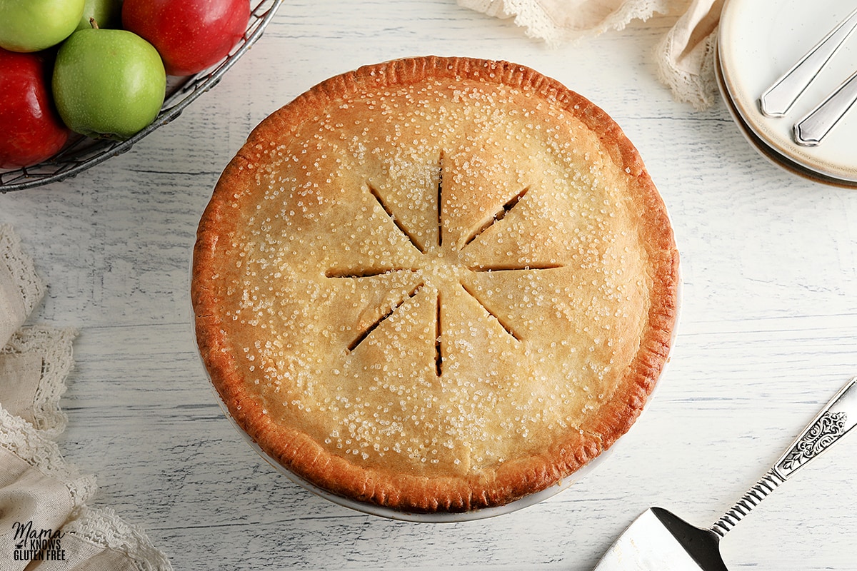 gluten-free apple pie with pie cutter, basket of apples, and white plates