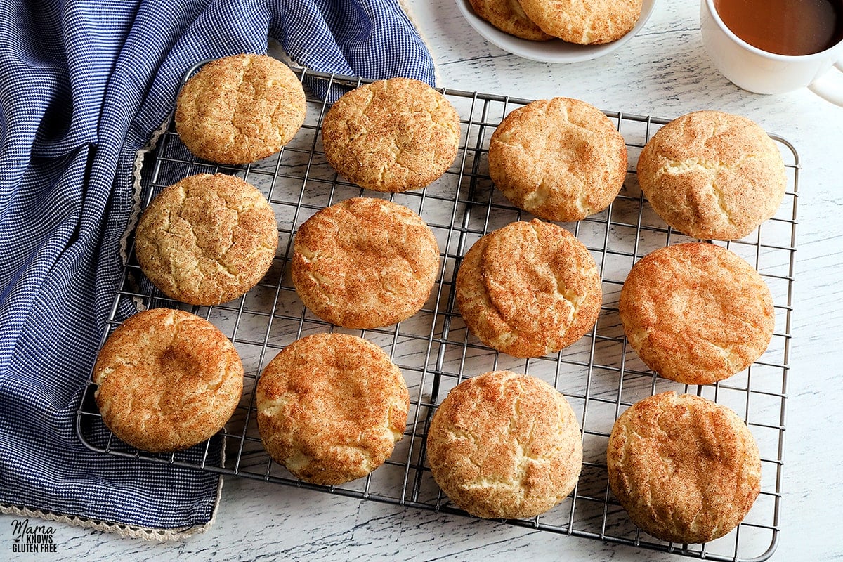 gluten-free snickerdoodle cookies on a sliver cooling rack with a plate of cookies, coffee, and a blue napkin in the background