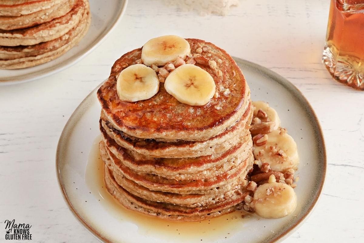 banana oatmeal pancakes on a white plate topped with sliced bananas, pecans, and syrup.