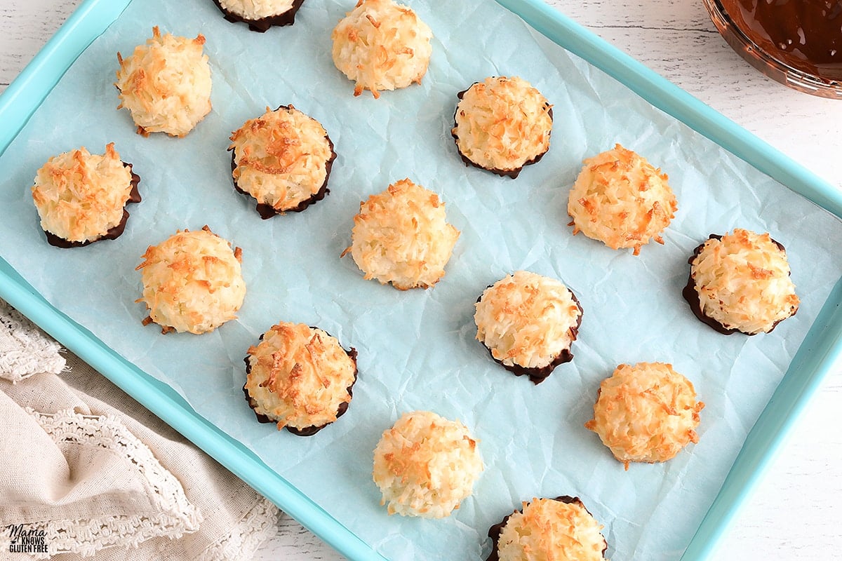 gluten-free macaroon cookies on a blue cookie sheet with a bowl of melted chocolate and kitchen towel in the background 