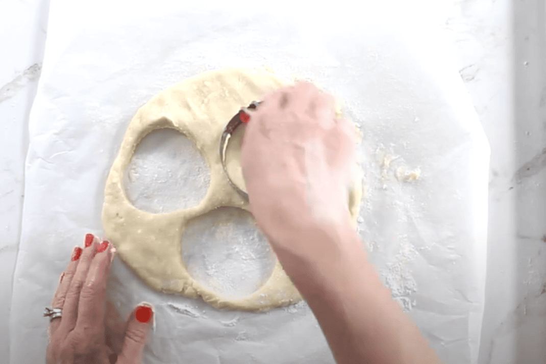 A hand presses a round biscuit cutter into rolled-out Danish dough to cut clean circles, with several rounds already removed from the dough on a floured sheet of parchment paper.