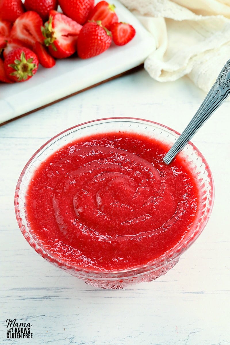 strawberry reduction sauce in a glass bowl with a silver spoon and strawberries and a cream napkin in the background