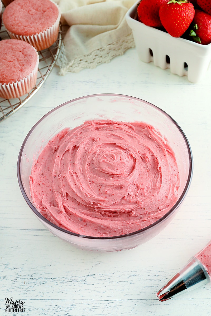 strawberry frosting in a glass bowl with cupcakes and strawberries in the background