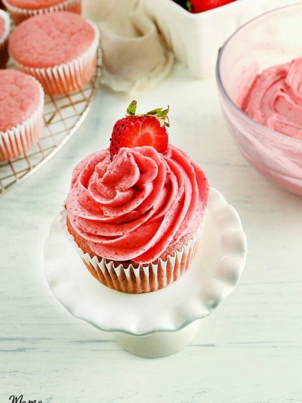 strawberry frosting piped on a strawberry cupcakes with bowl of frosting, strawberries, and cupcakes in the background