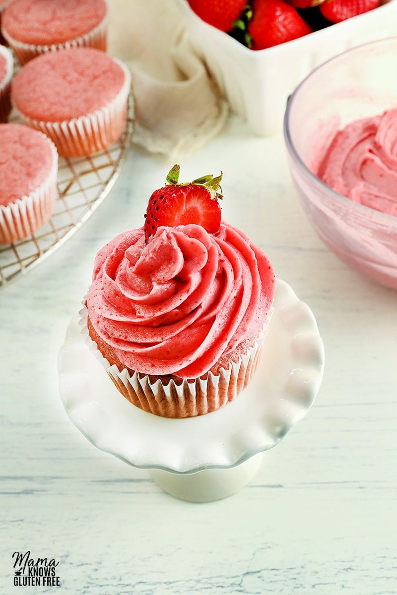 strawberry frosting piped on a strawberry cupcakes with bowl of frosting, strawberries, and cupcakes in the background