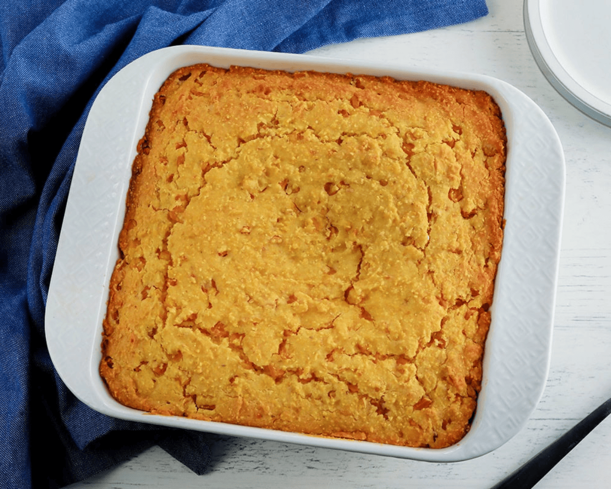 Golden baked gluten-free corn casserole in a white square baking dish, resting on a blue cloth with a lightly cracked, tender top.