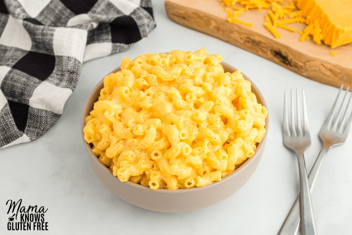 gluten-free mac and cheese in a bowl with two forks, napkin, and cutting board with cheese in the background
