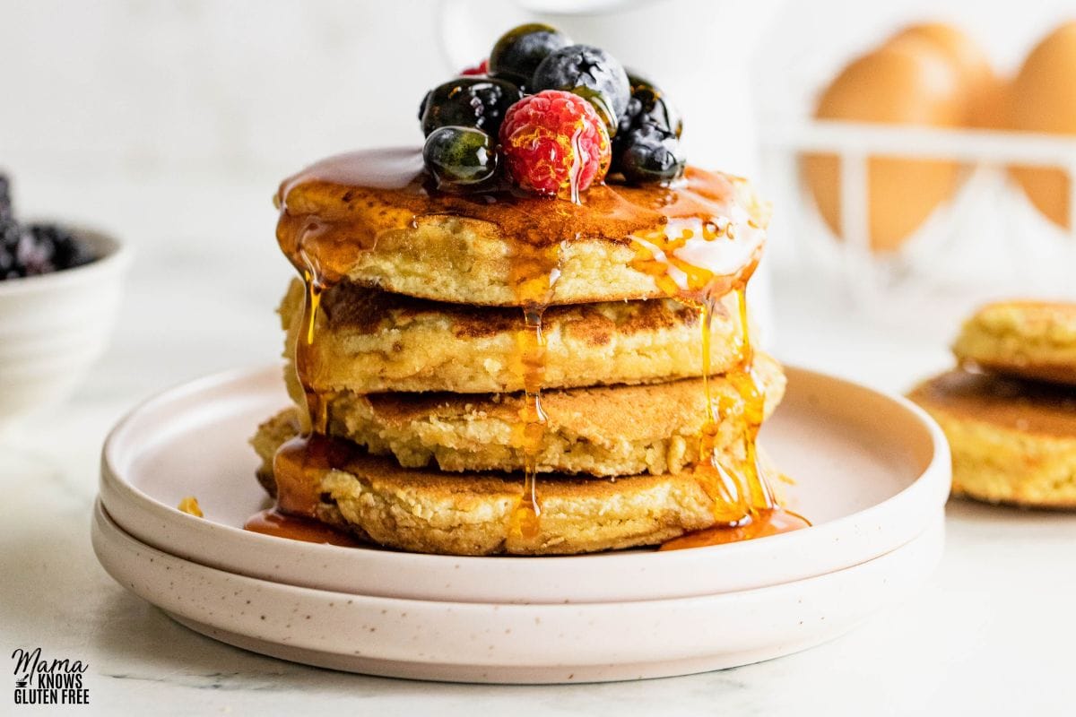 A stack of almond flour pancakes with berries and syrup on top. Eggs in the background.